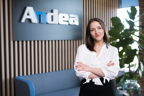 A woman stands in an office, arms crossed, in front of a dark blue sign with the white and blue Andes logo. She wears a white blouse and black skirt, with red nail polish. A large green plant is to her right. This represents innovation in manufacturing or digital transformation.