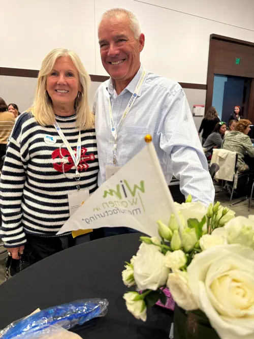 A smiling woman in a striped sweater and a man in a checkered shirt stand together at tables with white roses and a WIM flag. They are at the WIM Summit.