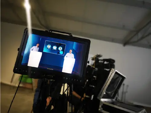 A monitor displays a production monitoring interface with two presenters on screen. A camera is set up in a studio environment for broadcasting. This highlights digital transformation in manufacturing.