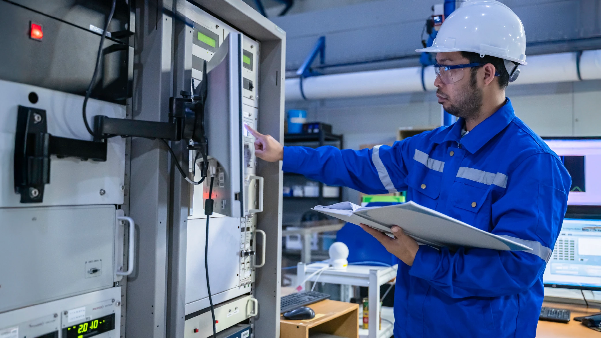 An engineer in a blue jumpsuit and hard hat studies a document while pressing a button on complex industrial electrical equipment. This visual represents manufacturing operations and digital transformation solutions.